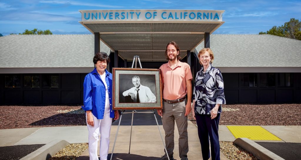 Thomas Scott standing in front of a building at the University of California with two women and a photograph of a man