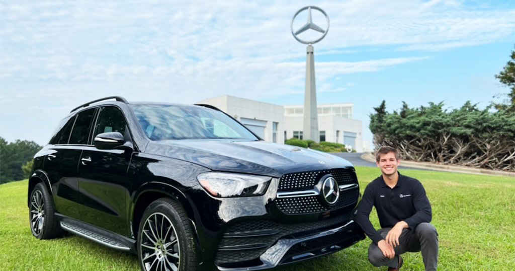 Blake Z at squatting in a field in front of the Mercedes building next to a vehicle